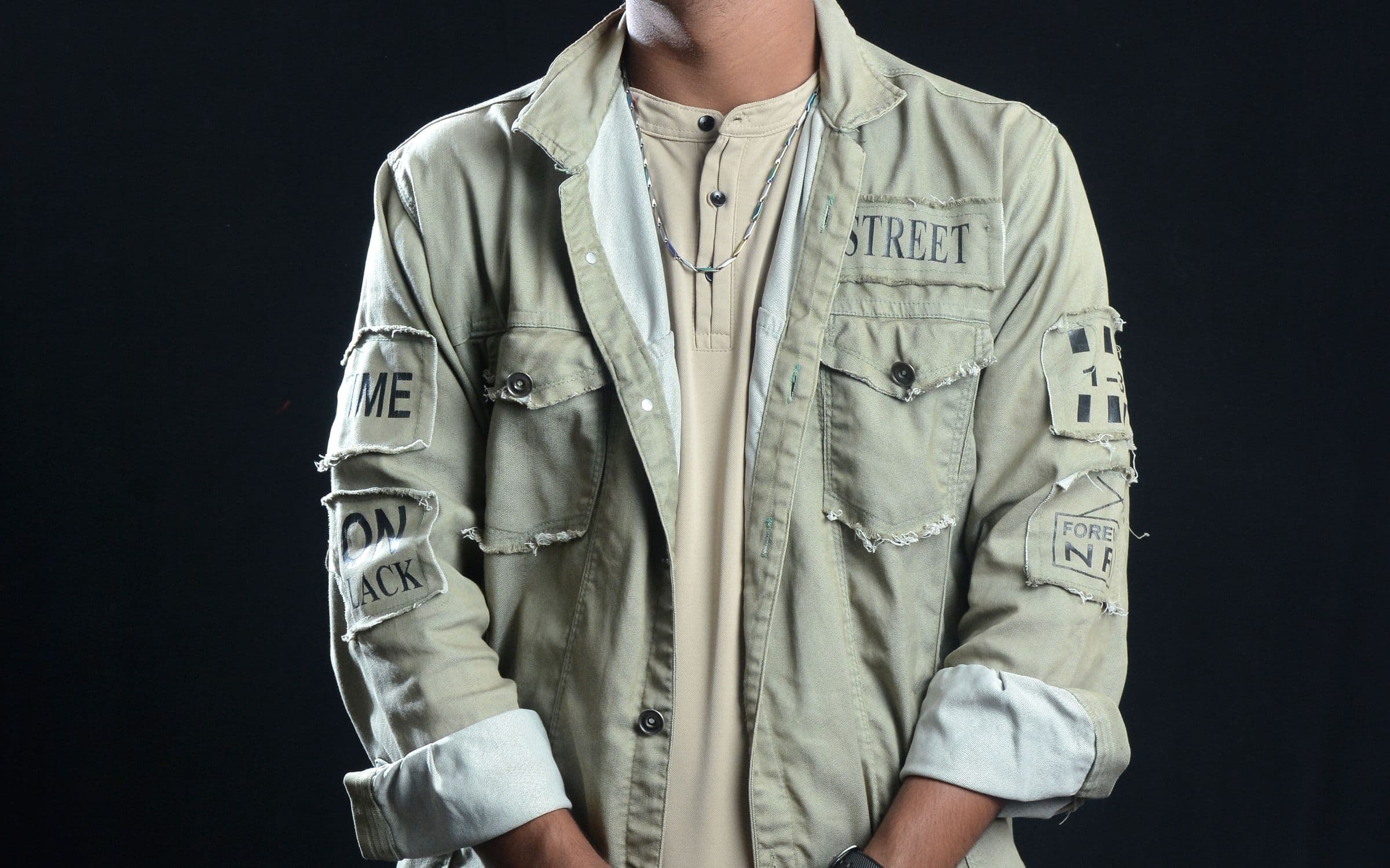 Ecommerce apparel shot: a model in a khaki utility jacket with frayed cloth patches, photographed against a black backdrop.