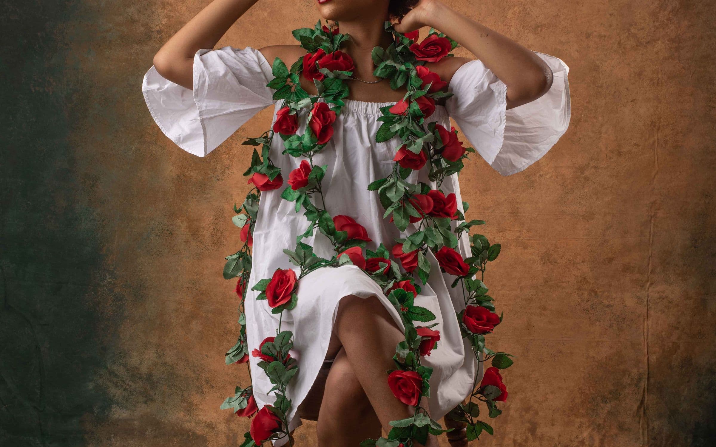 Ecommerce fashion shot: a model in a white off-the-shoulder dress draped with a garland of red roses, against a hand-painted backdrop.